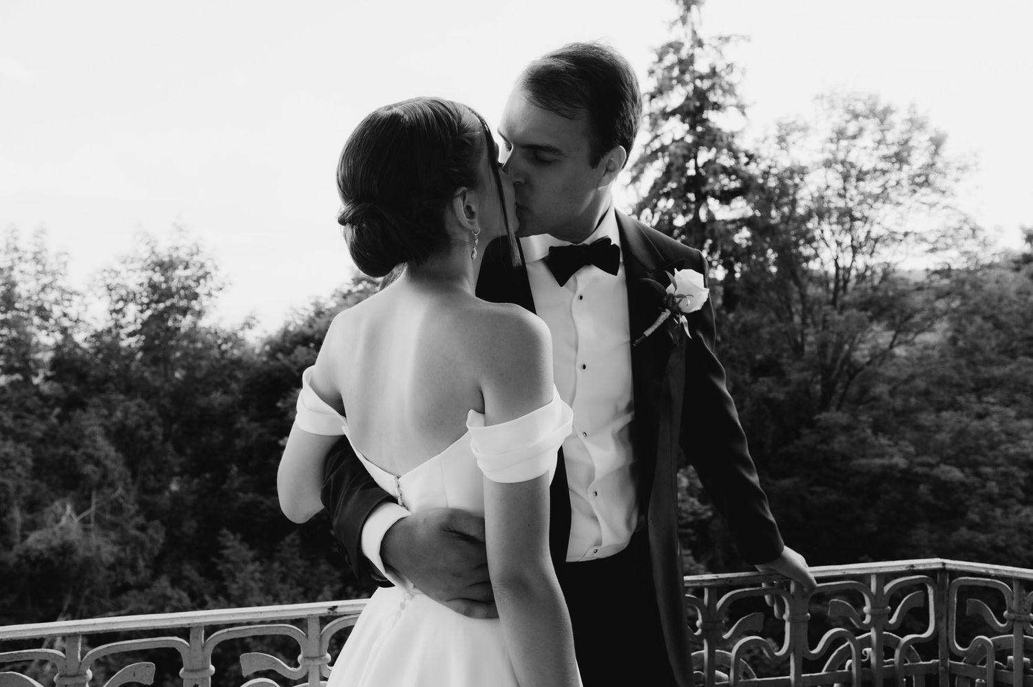 Black and white wedding photo of bride and groom sharing a romantic kiss on a balcony surrounded by lush trees.
