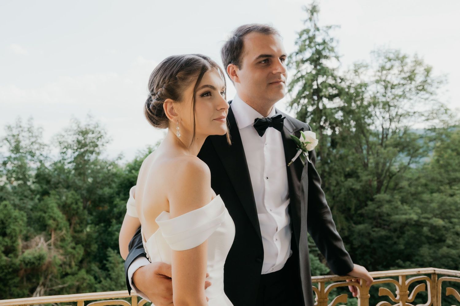 Bride and groom in wedding attire pose on ornate balcony surrounded by lush green trees.