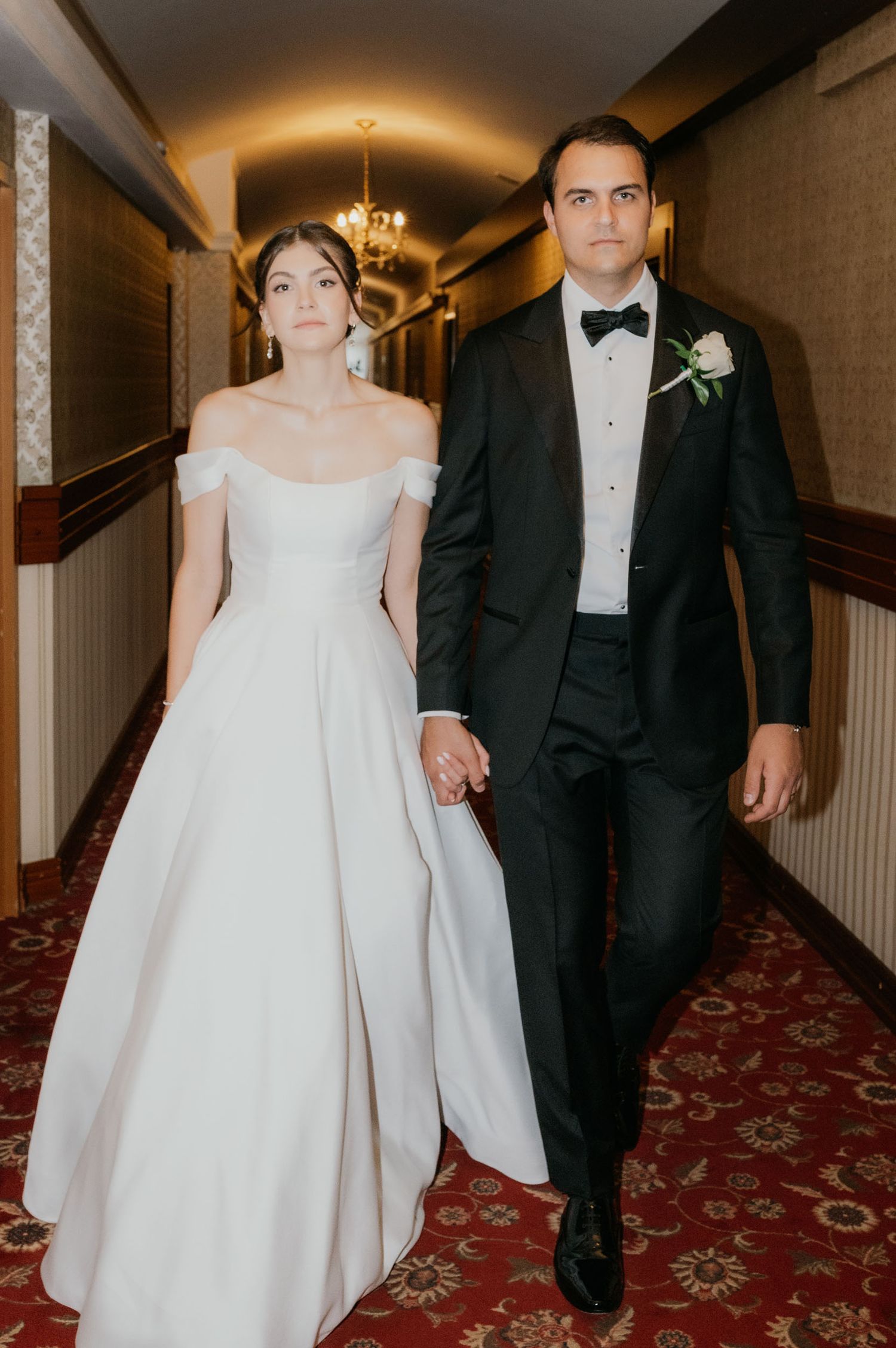 Bride in off-shoulder white gown and groom in black tuxedo walk hand in hand down an elegant hotel hallway.