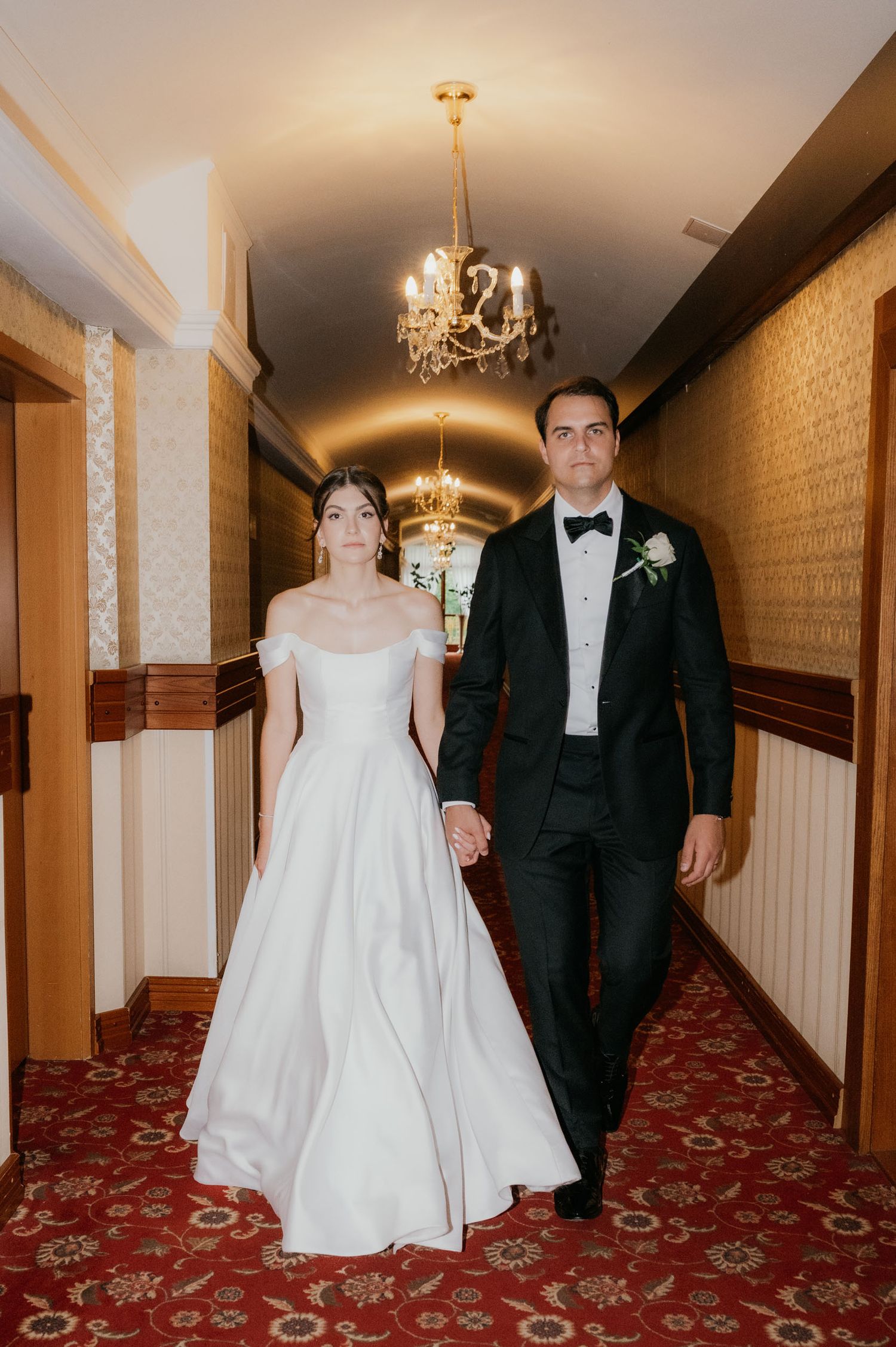A bride in a white off-shoulder gown and groom in black tuxedo walk hand-in-hand through an elegant chandelier-lit hallway.
