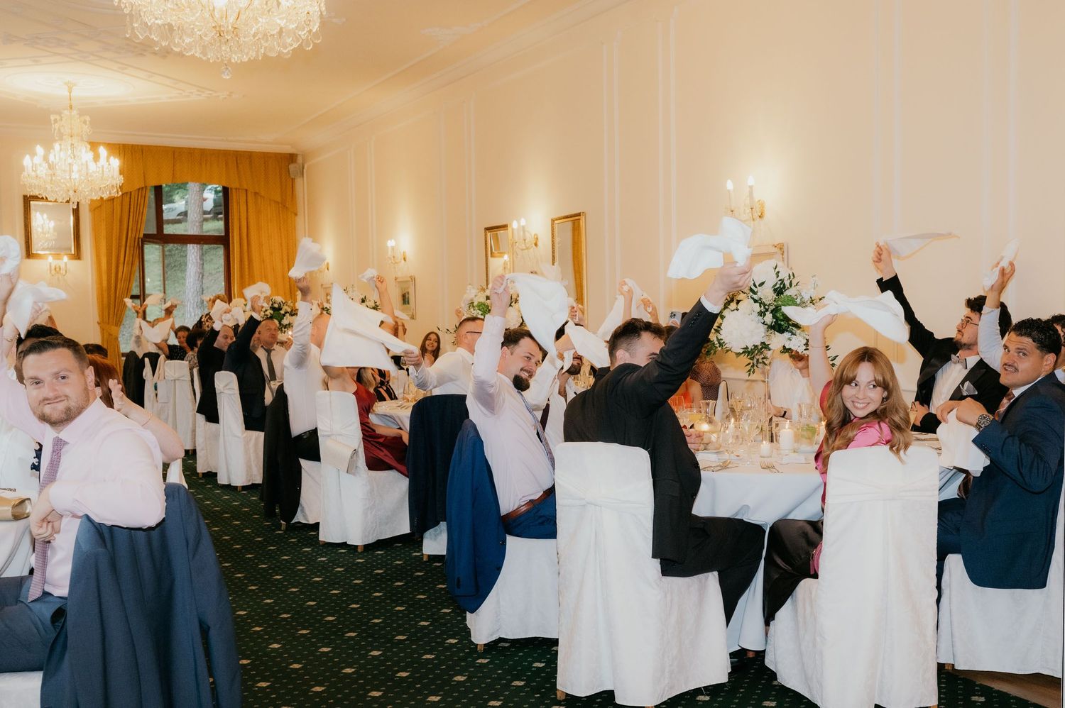 Wedding guests raising white feather fans in elegant ballroom with chandeliers and white chair covers.