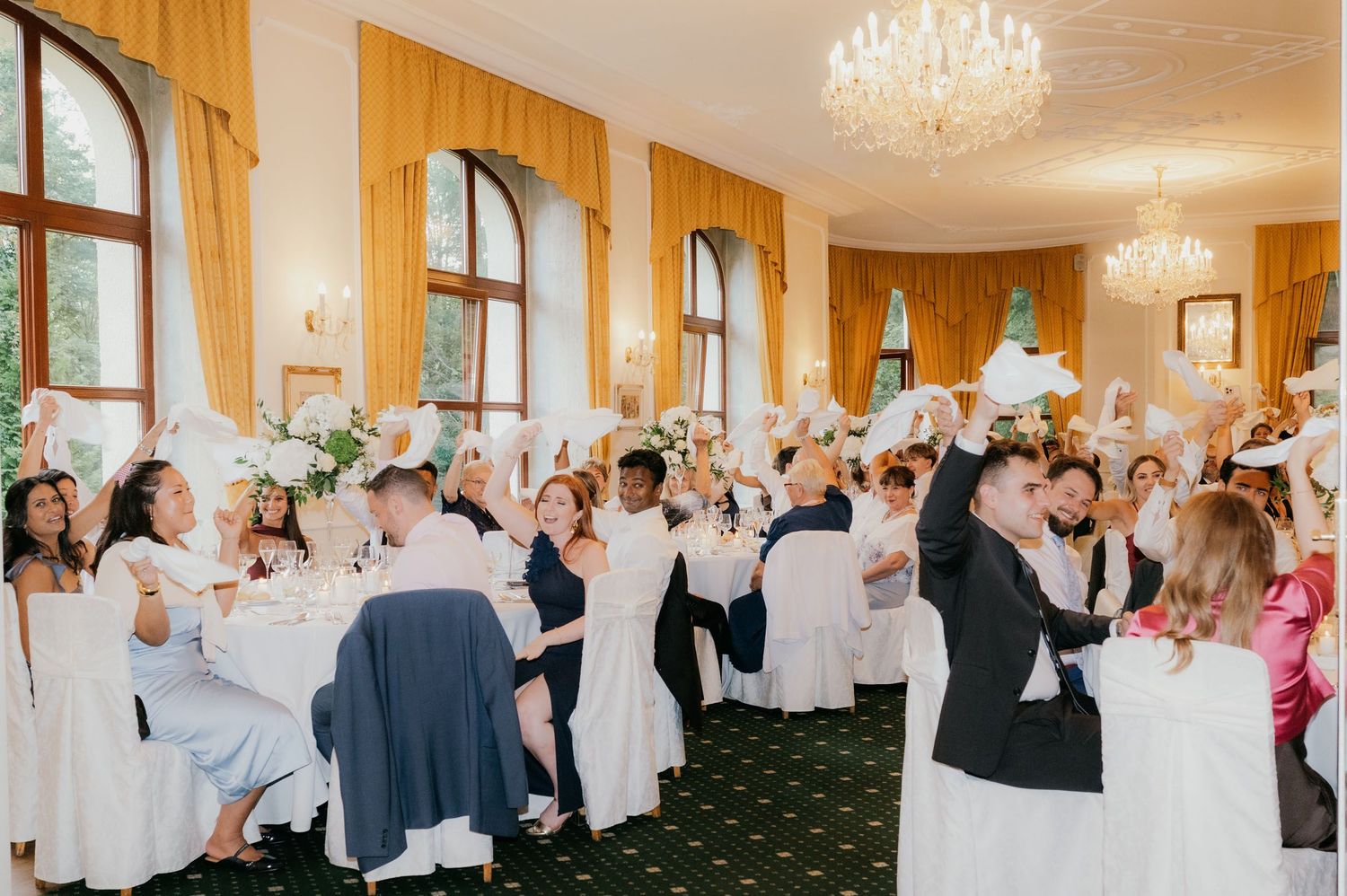 Elegant wedding reception in a grand ballroom with guests raising white napkins in celebration.