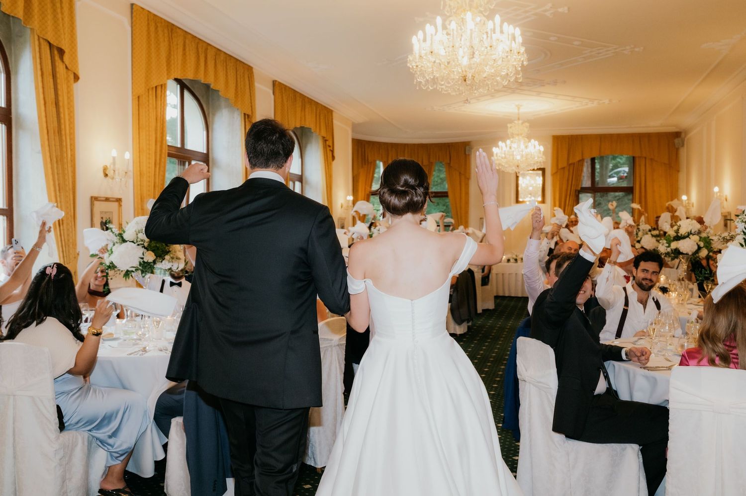 Bride and groom enter elegant wedding reception hall with cheering guests at decorated white tables under chandelier.