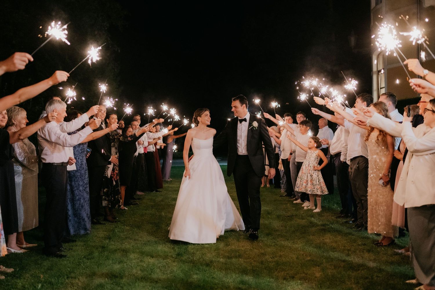 Bride and groom walk through sparkler exit surrounded by cheering wedding guests at night.
