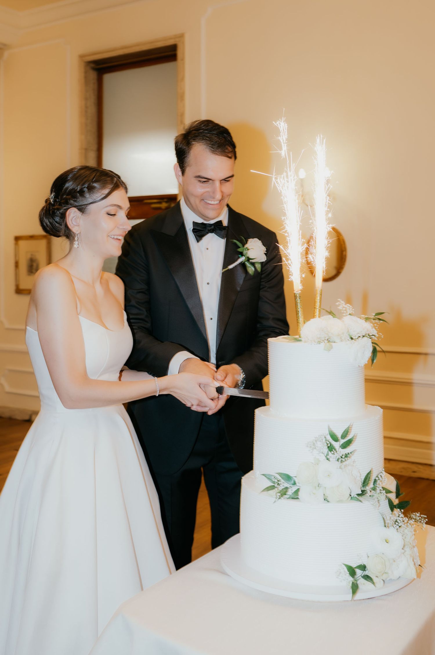 Bride and groom cutting a white three-tier wedding cake decorated with greenery and sparklers.