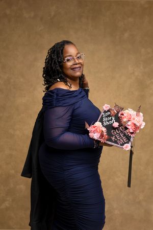 A smiling woman in a navy blue off-shoulder dress holds a decorated graduation cap with pink roses at a photo studio.