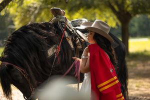 Woman in red cape and white cowboy hat stands beside a large black horse in a sunlit outdoor setting.