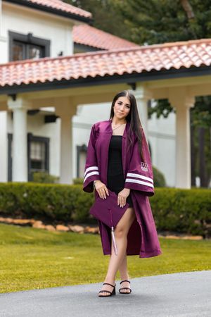 Young woman in maroon graduation cap and gown posing outdoors in front of elegant building with red tile roof.