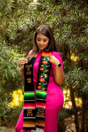 Young Latina woman in pink dress holding Class of 2025 graduation stole with colorful Mexican embroidery outdoors.