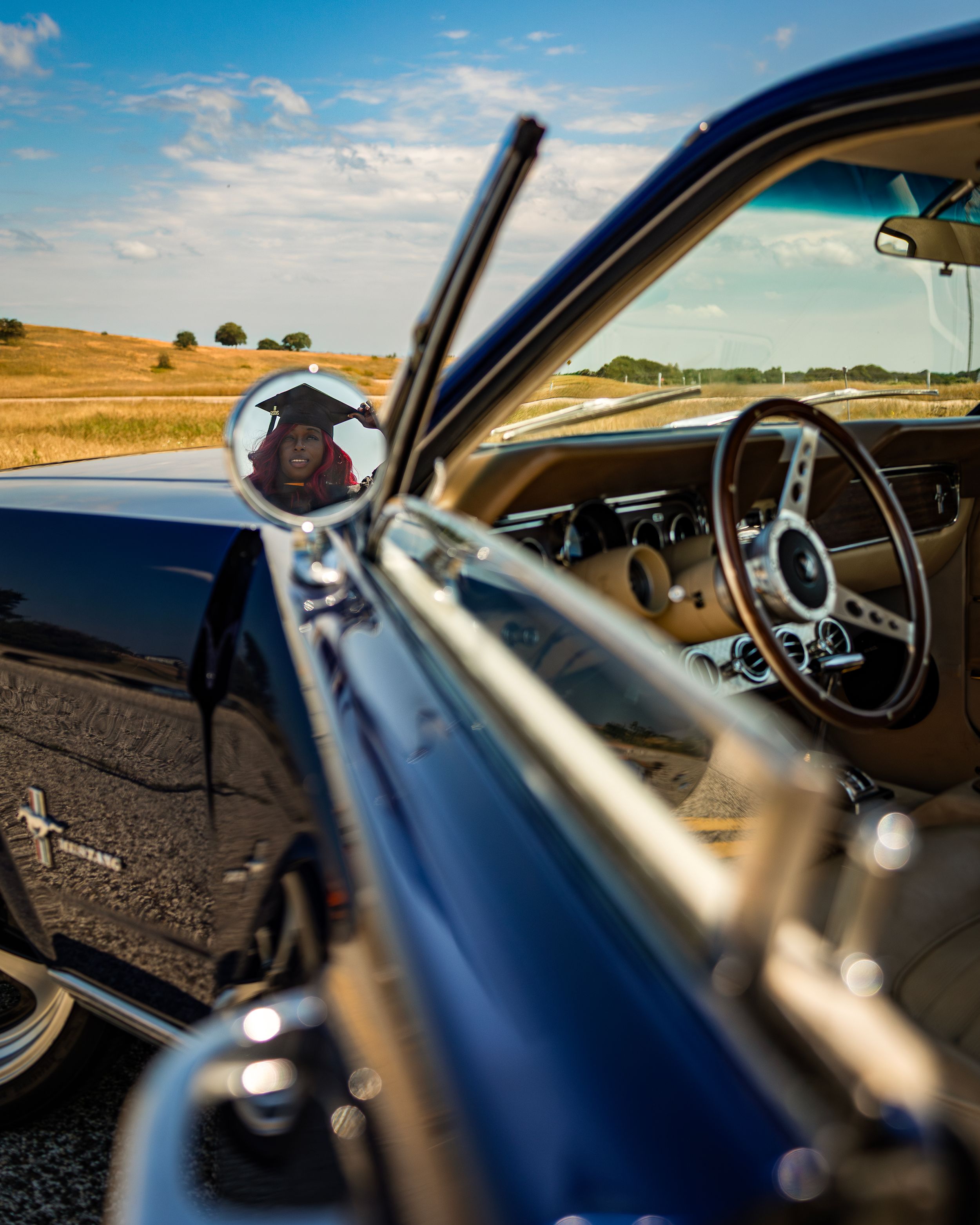 Classic blue muscle car interior with steering wheel and side mirror reflecting a person, parked on a rural road under blue skies.