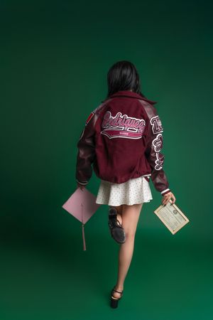 Young woman in burgundy varsity jacket, white skirt, holding graduation cap and diploma against green background.