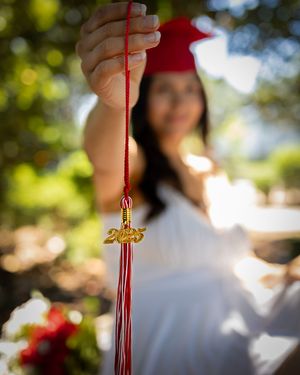 A graduate in red cap holds a 2025 gold tassel charm on a red cord outdoors.