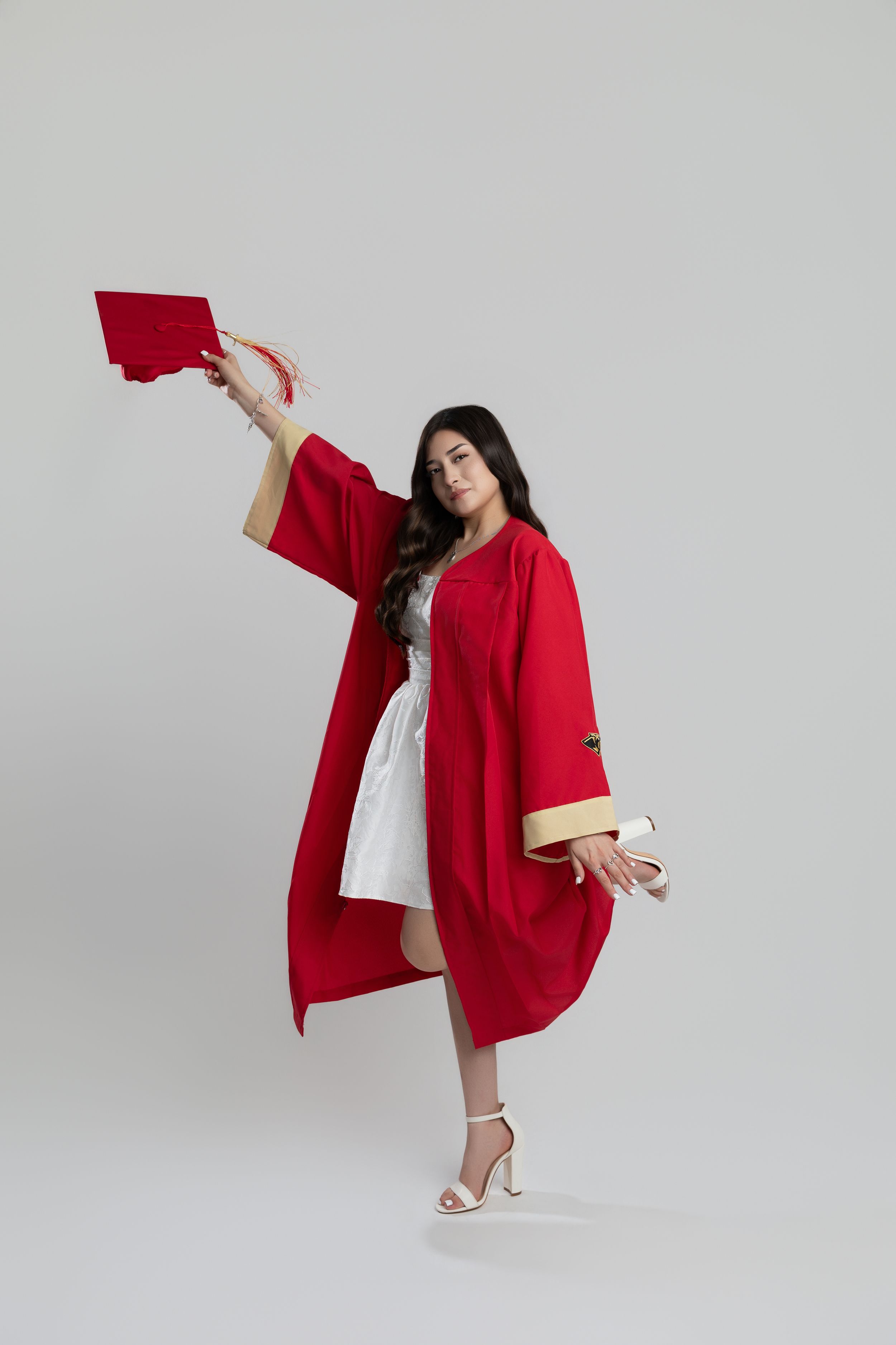 Young woman in red graduation gown and cap celebrating outdoors, posing dynamically in white dress and heels.
