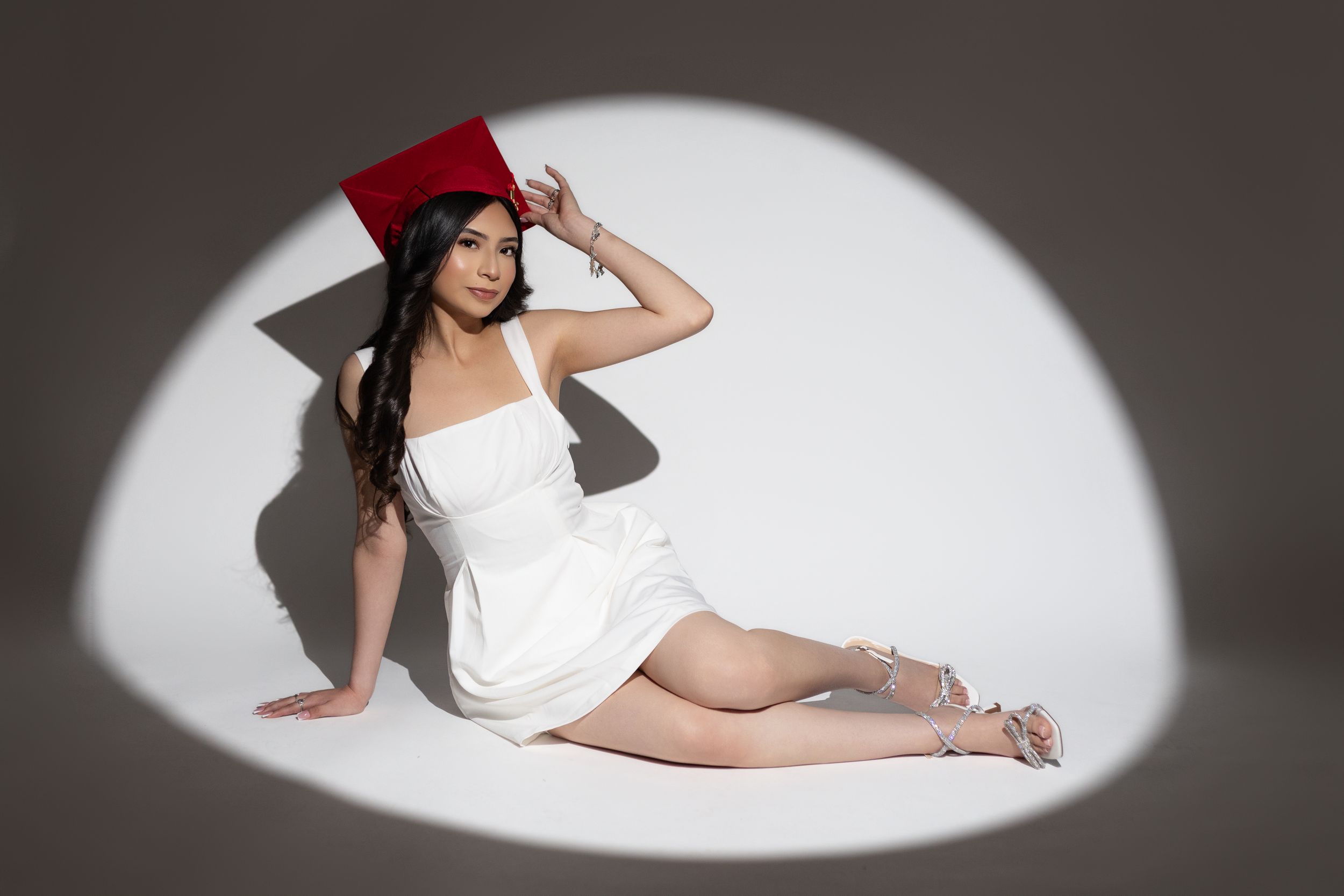 Young woman in white mini dress and red graduation cap poses gracefully on floor in dramatic spotlight.