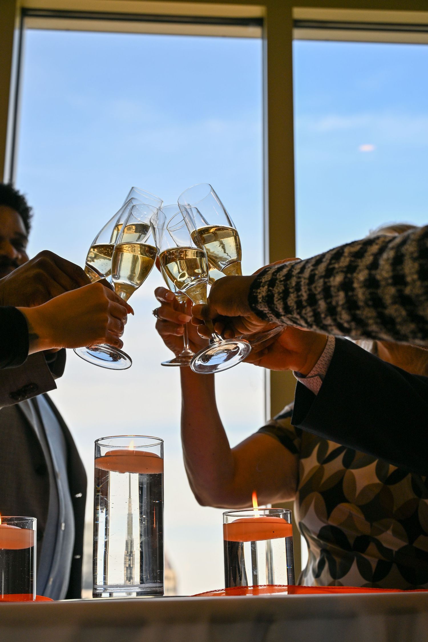 Group of people toasting with champagne glasses near a sunlit window with candles on the table.