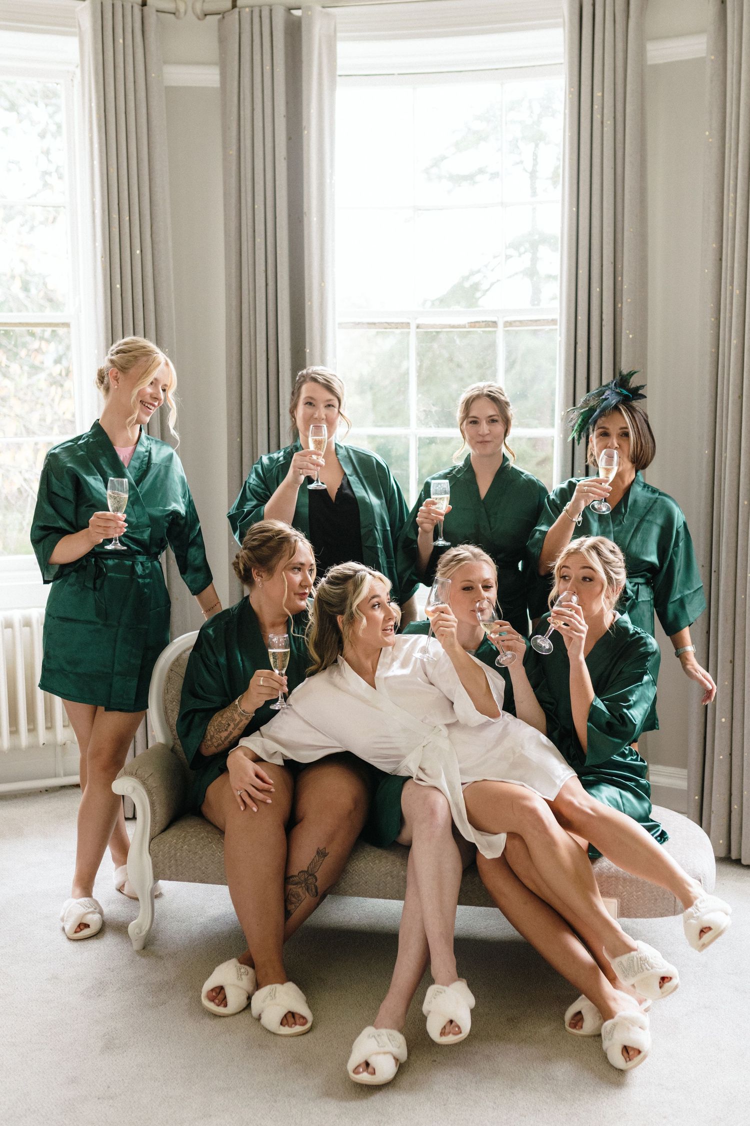 Bride in white robe with bridesmaids in green satin robes toasting champagne glasses before wedding ceremony.
