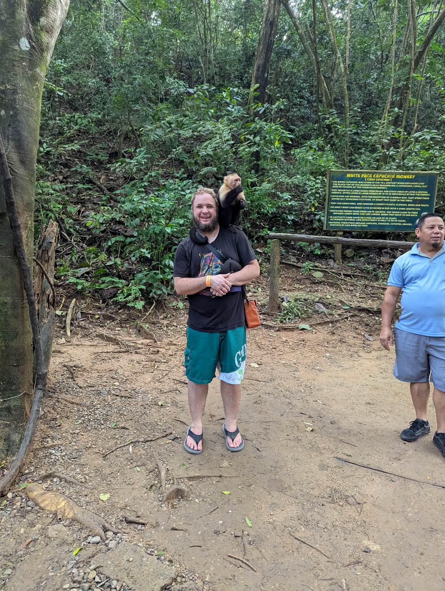 Man in black shirt and green shorts stands on dirt trail surrounded by lush tropical forest near an informational sign.