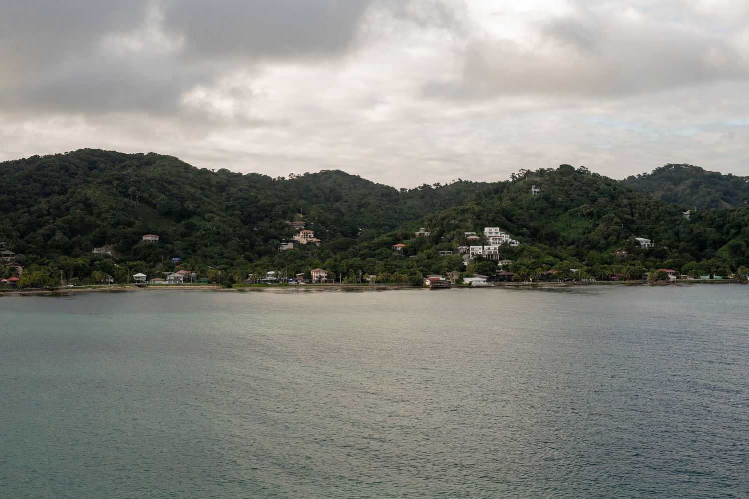 Lush green hillside tropical town with white buildings overlooking calm turquoise bay waters under cloudy skies.