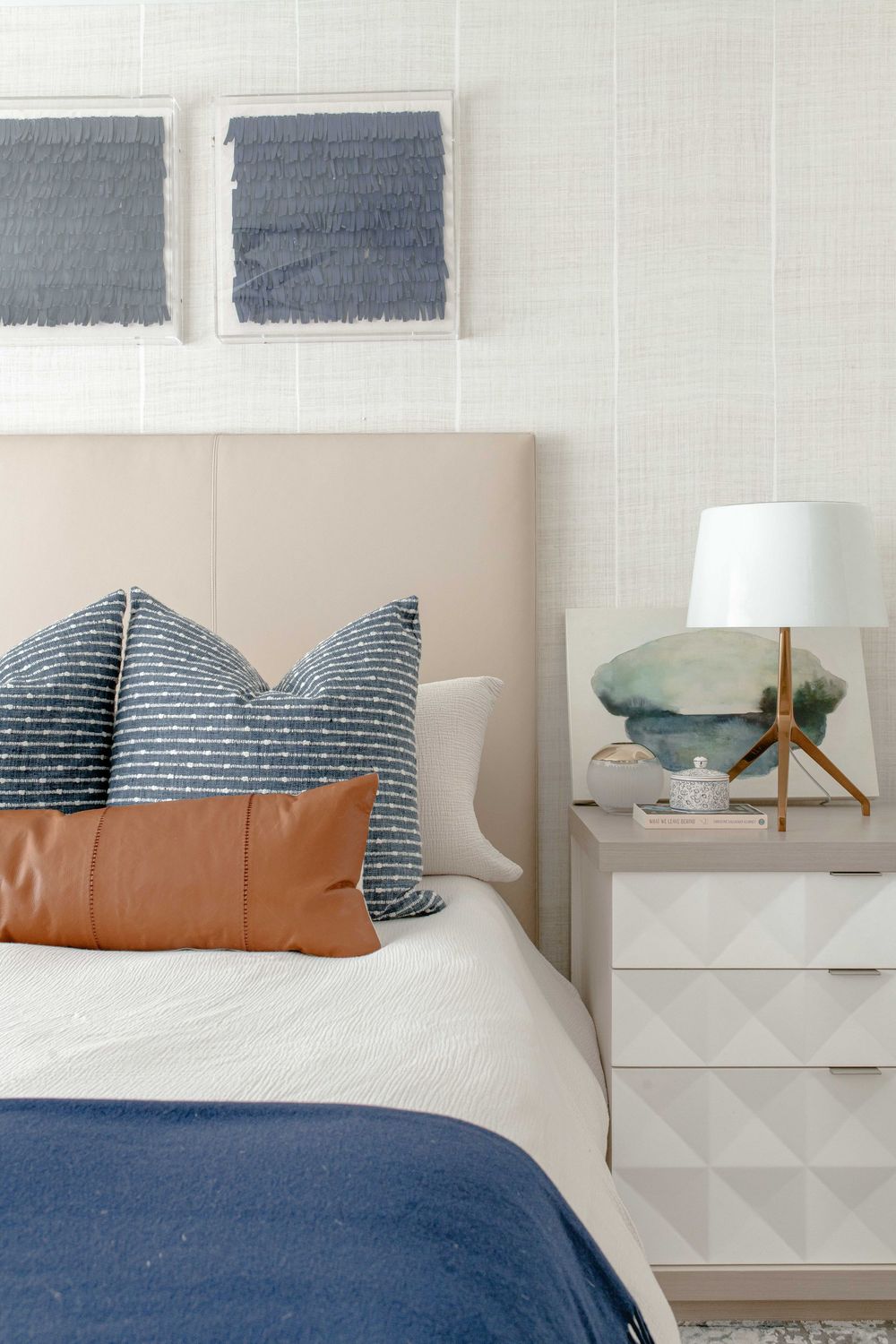Neutral bedroom with beige upholstered headboard, navy blue pillows, leather lumbar pillow, and geometric nightstand.