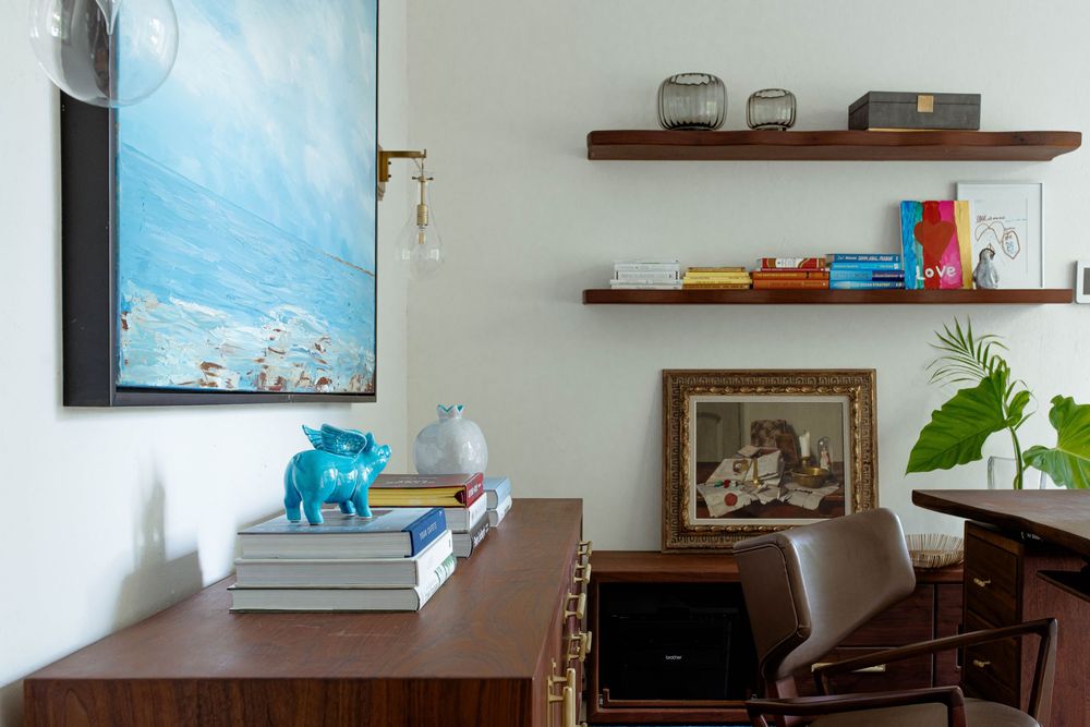 Modern home office with walnut desk, blue art, floating shelves with books, and a vintage framed portrait.