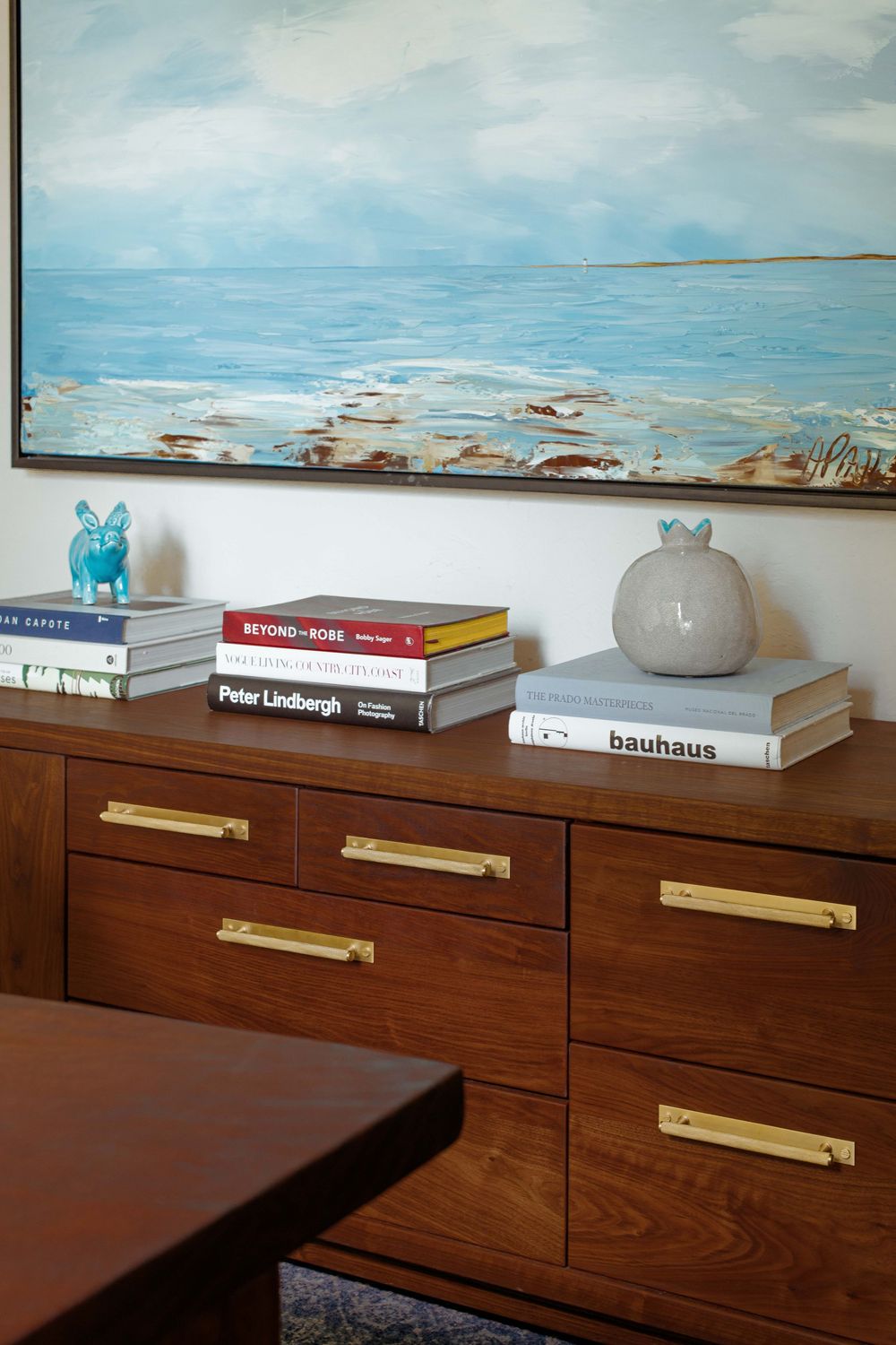 Mid-century modern walnut dresser with brass handles, stacked coffee table books, and coastal painting above.