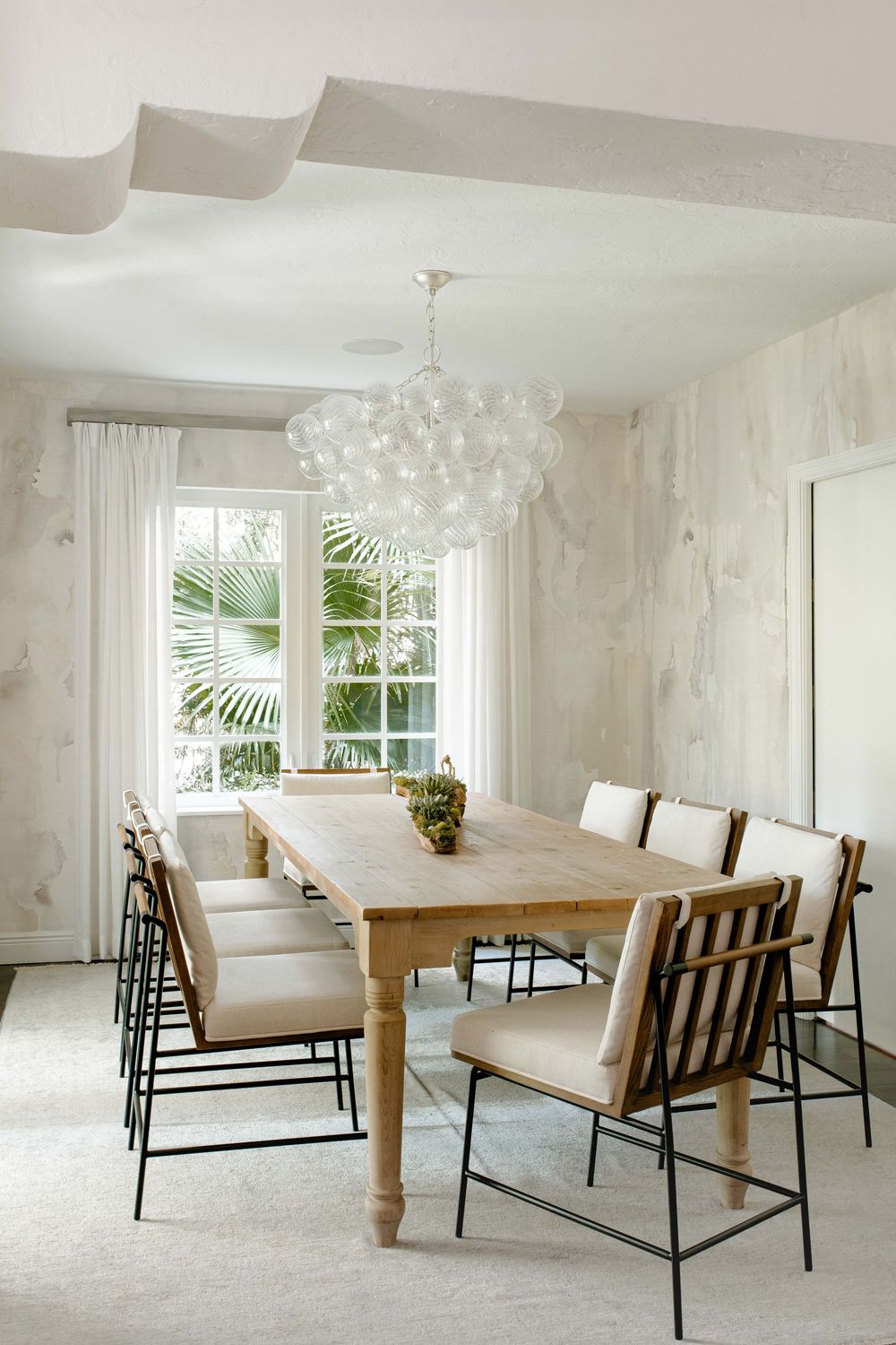 Airy dining room with rustic wood table, mixed chairs, bubble glass chandelier, marble walls, and tropical window view.