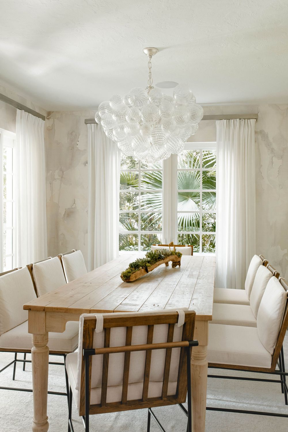 Elegant dining room with long wooden table, white upholstered chairs, bubble glass chandelier, and tropical garden view.