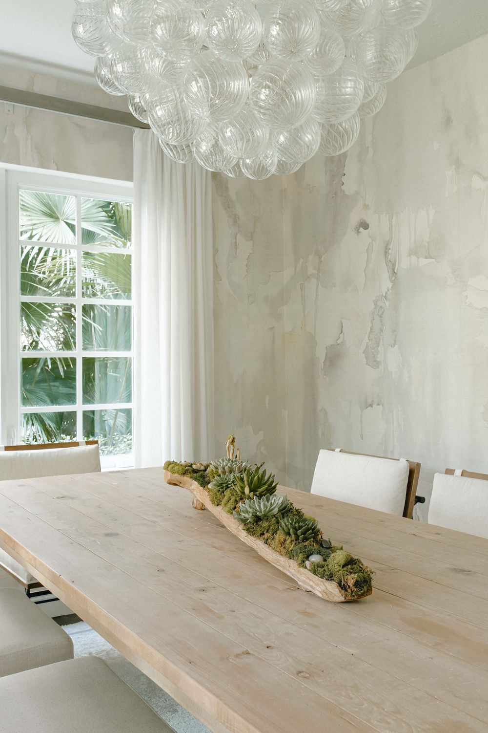 Close-up of light wood dining table with succulent centerpiece under bubble chandelier with marble accent wall.