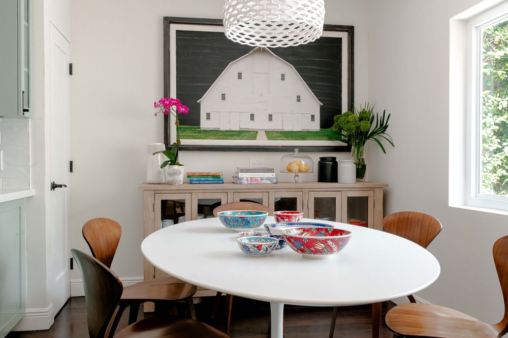 Modern dining room with white oval table, wooden chairs, barn painting, and colorful bowls on a globe pendant light.