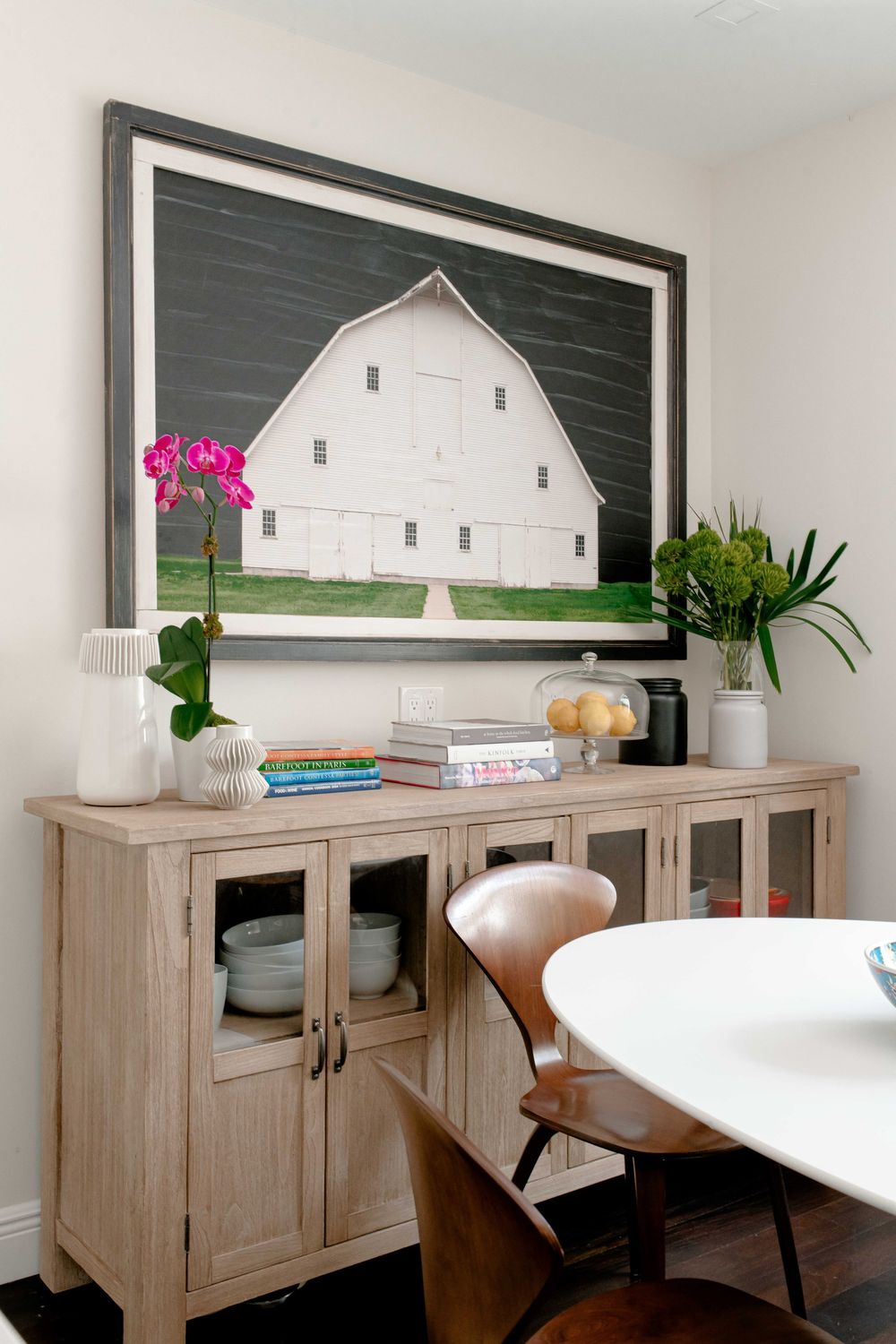 Dining area with a light wood buffet, large barn photograph, orchid, and modern white table with leather chairs.