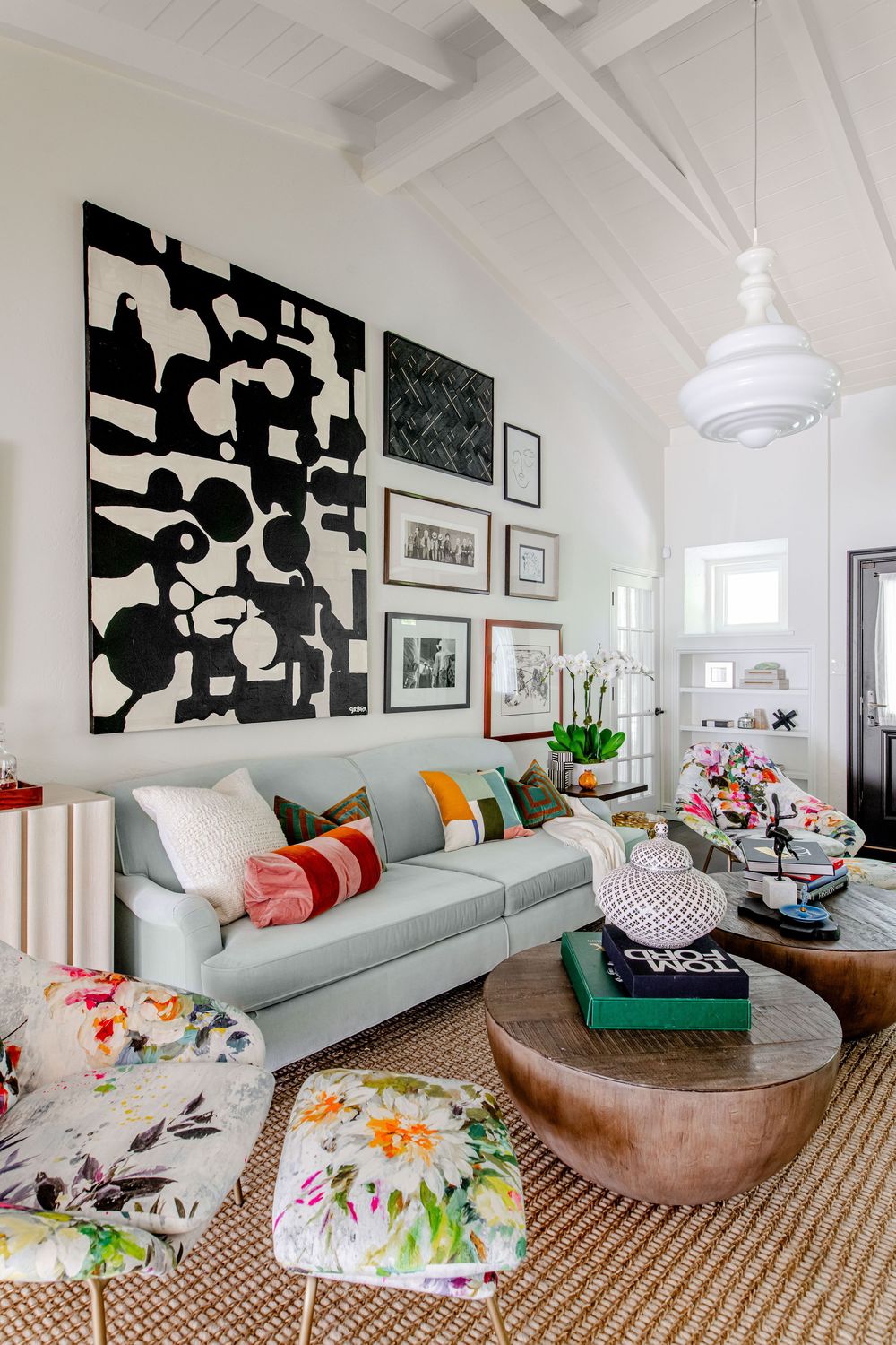 Eclectic living room with light blue sofa, colorful floral ottomans, bold black and white wall art, and wooden coffee table.