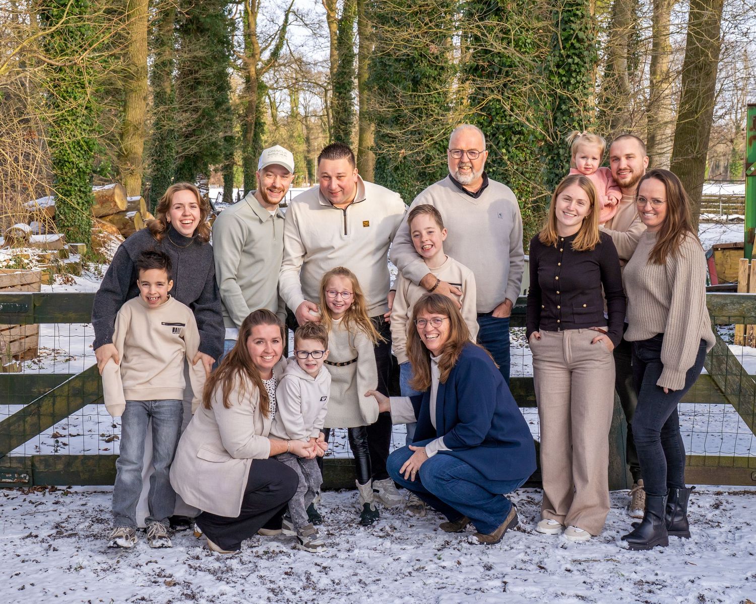 Een grote familiegroep poseert buiten in een besneeuwde winteromgeving, vlakbij een houten hek omringd door met sneeuw bedekte bomen.