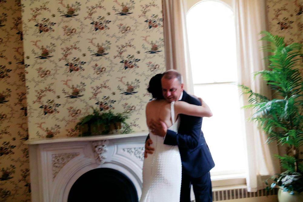 Bride and father sharing a tender embrace near a fireplace in an elegant room with floral wallpaper.