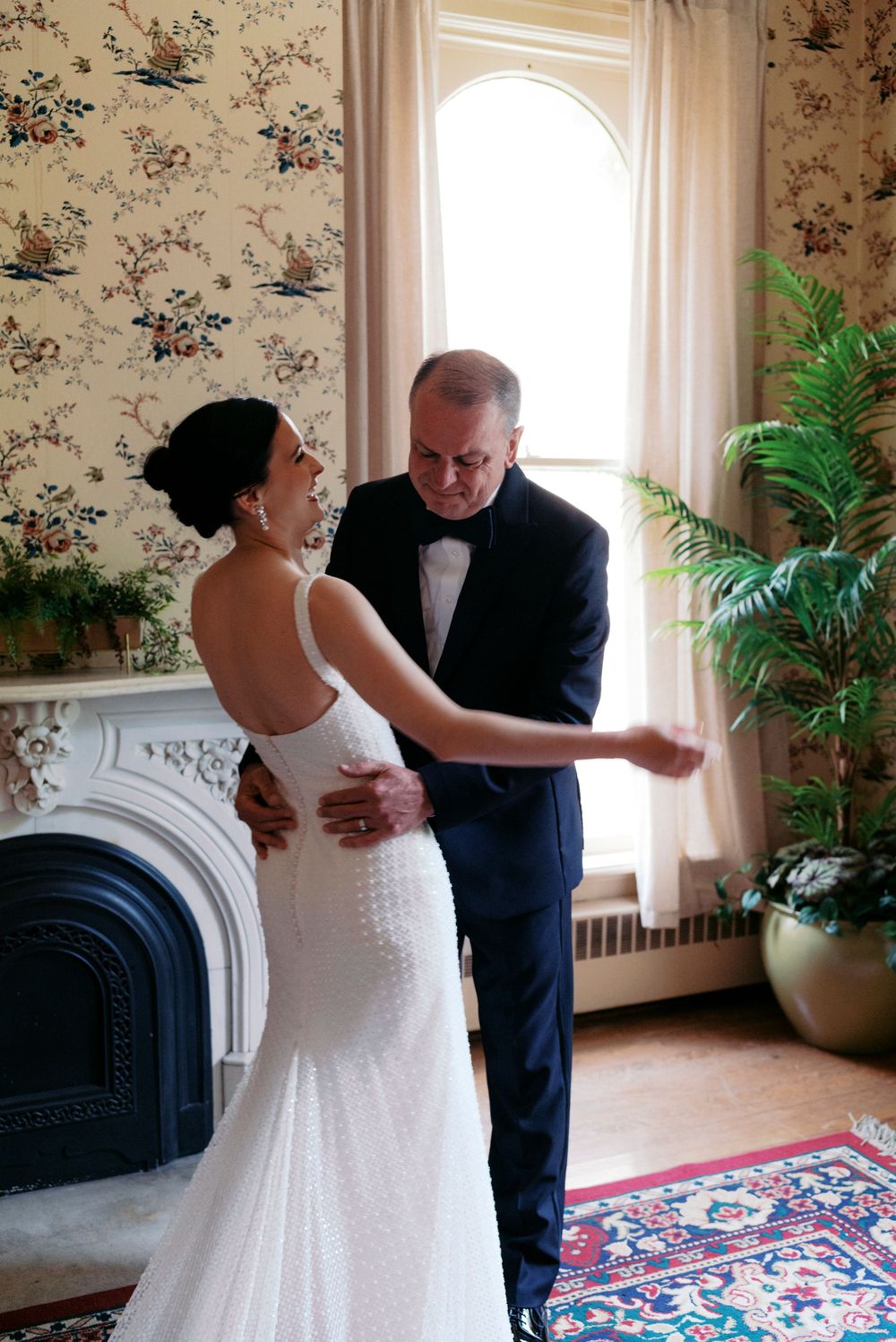 Midwest bride in white gown and father in tuxedo share a tender moment by a fireplace in an elegant floral room.