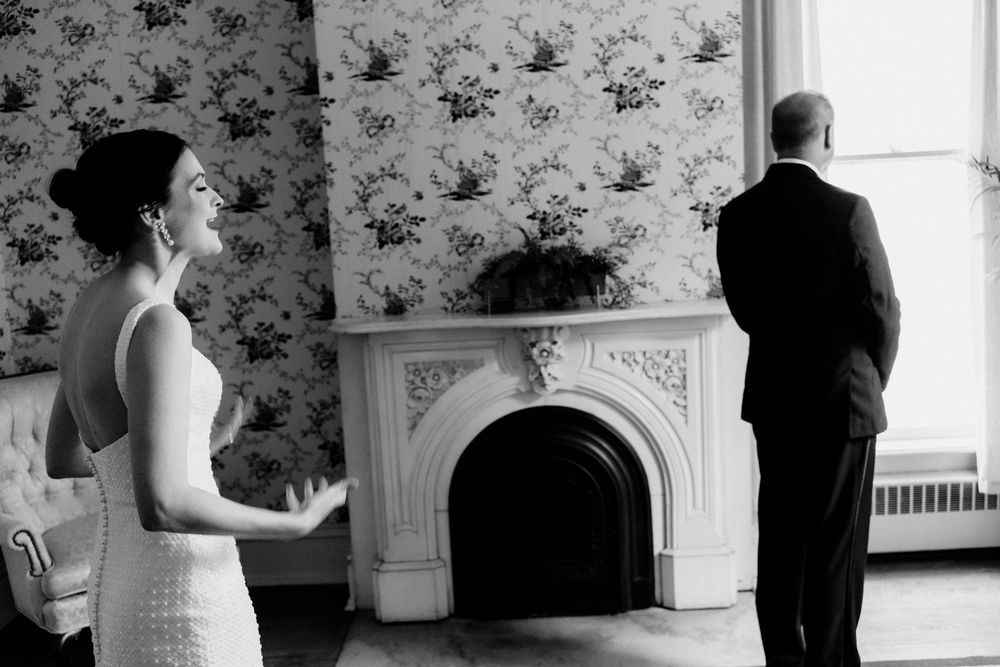 Intimate wedding photo of bride and father sharing a first look moment by an ornate historic fireplace.