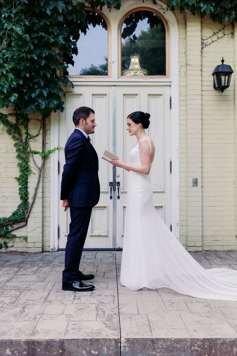Milwaukee bride reads vows to groom in navy suit outside yellow Victorian building with ivy and white doors.