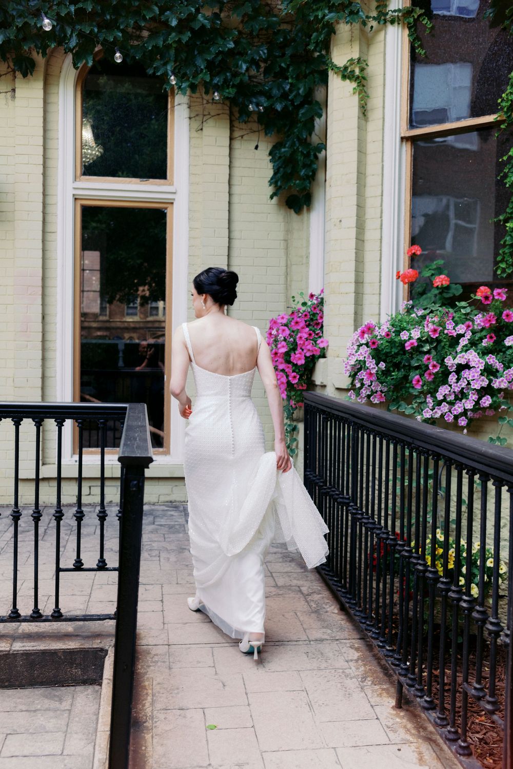 Bride in stylish white backless gown walks along a floral-adorned historic Milwaukee building entrance with lush pink flowers.