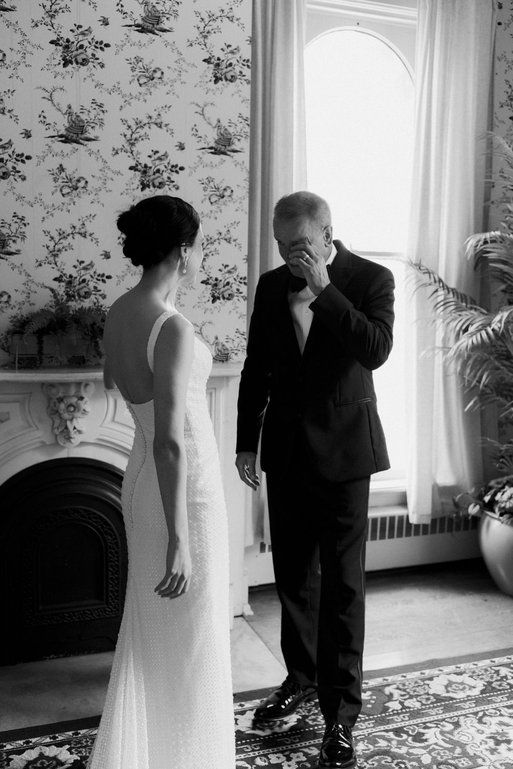 Black and white photo of emotional father wiping tears away as graceful bride in wedding gown turns away in ornate floral room.