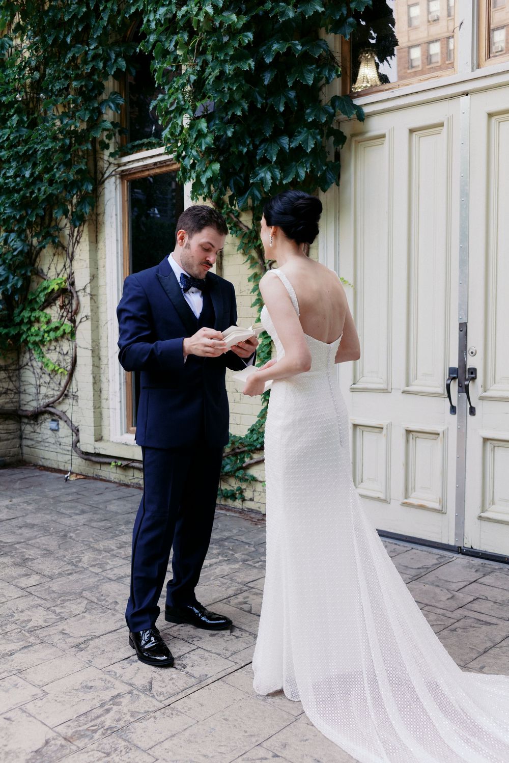 Groom in navy suit reads vows to bride in flowy white gown outside ivy-covered Milwaukee building during intimate vow ceremony.
