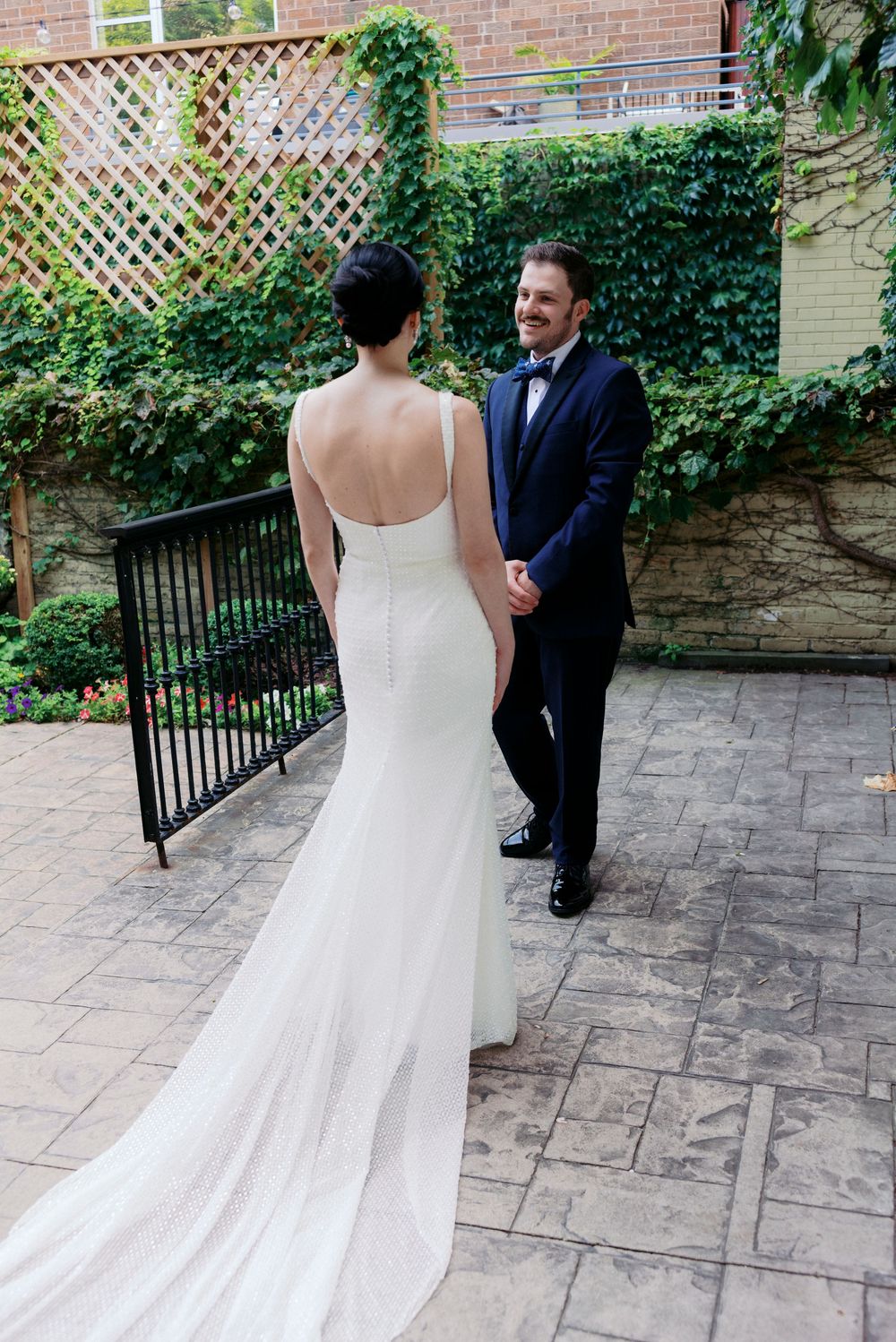 Bride in sophisticated backless white gown holds hands with groom in navy suit during outdoor first look ceremony.