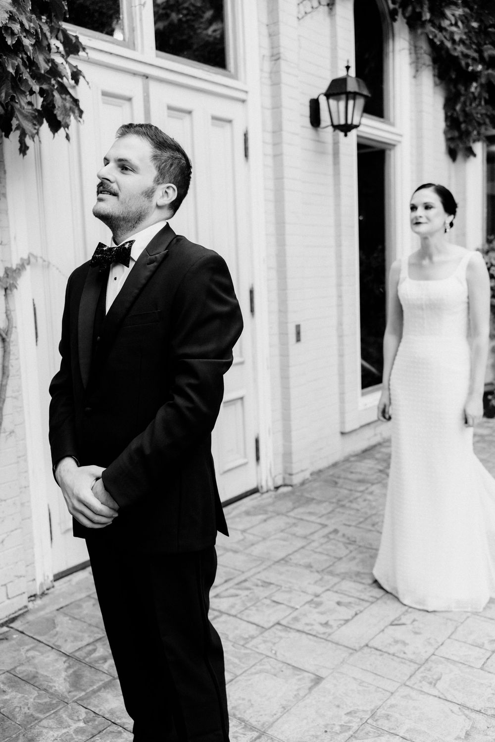 Black and white photo of a groom in a tuxedo standing outdoors as his bride in a white gown approaches behind him.