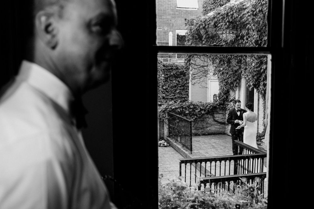 Documentary wedding photo of guest inside looking through rustic window at bride and groom standing on ivy-covered brick building steps.