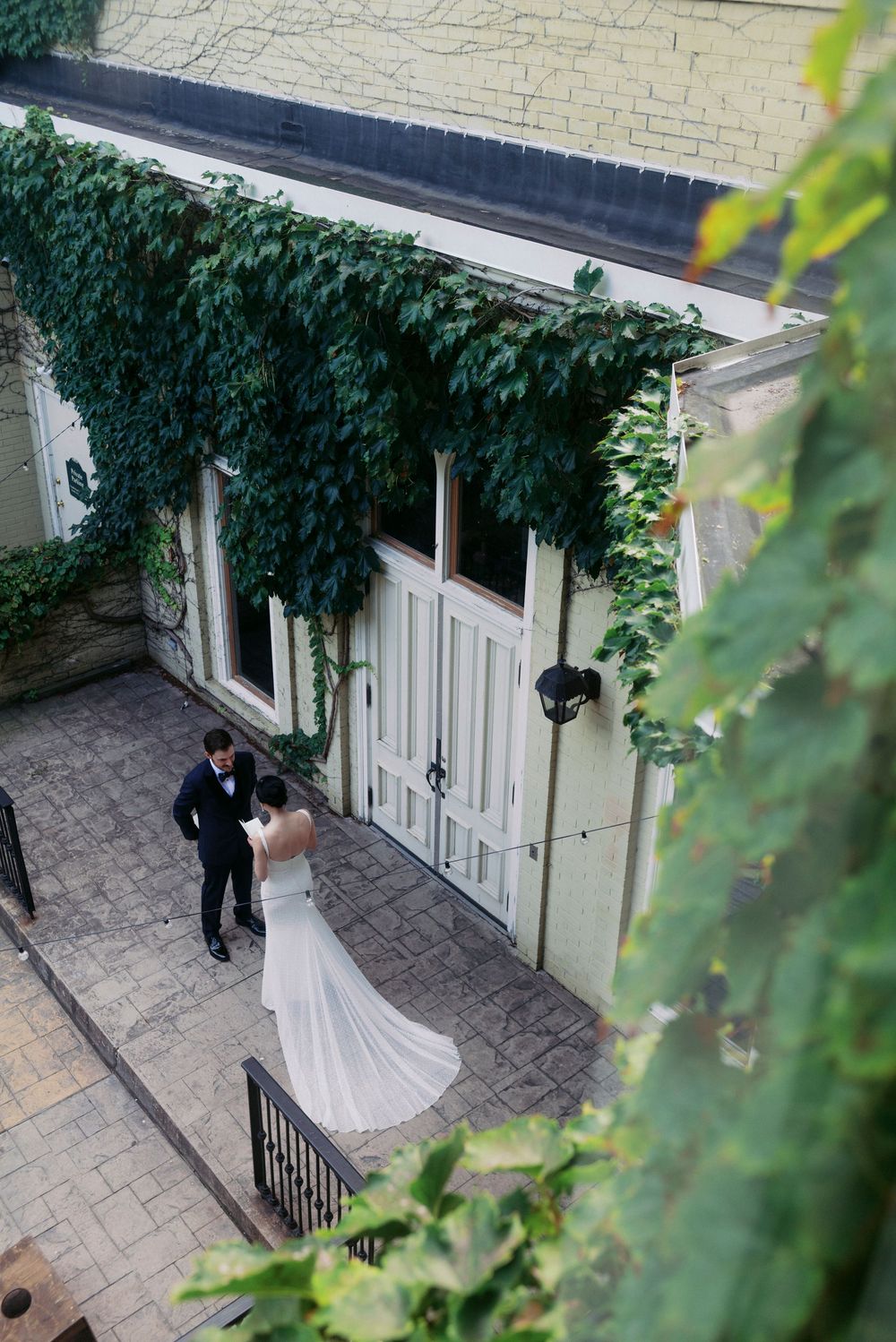 Aerial view of bride and groom sharing a romantic moment by ivy-covered white doors on a cobblestone courtyard in poetic moment.