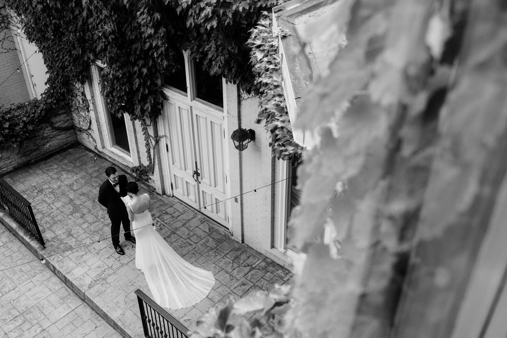 Black and white aerial photo of bride and groom standing together outside ivy-covered building on wedding day.