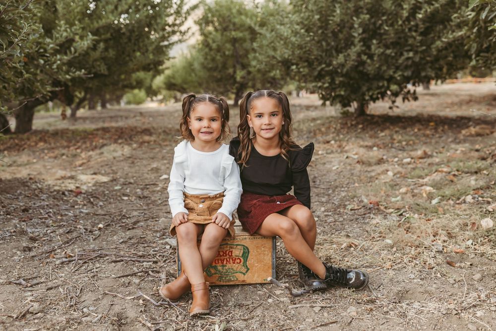 Two young sisters sit together on a vintage suitcase in an orchard, wearing stylish fall outfits.
