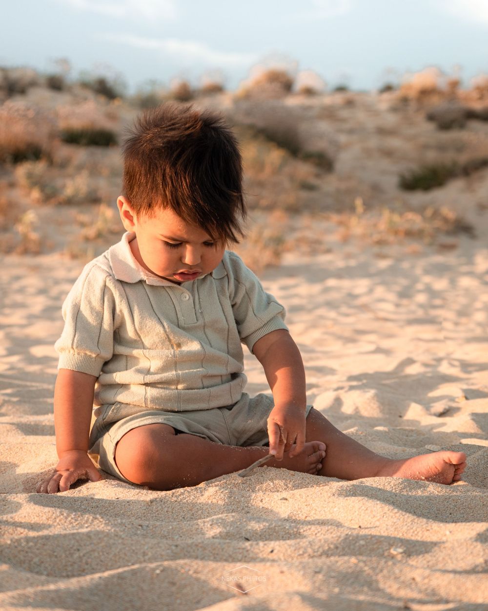 Adorable toddler boy sitting on sandy beach, playing with sand, wearing a light striped polo outfit.