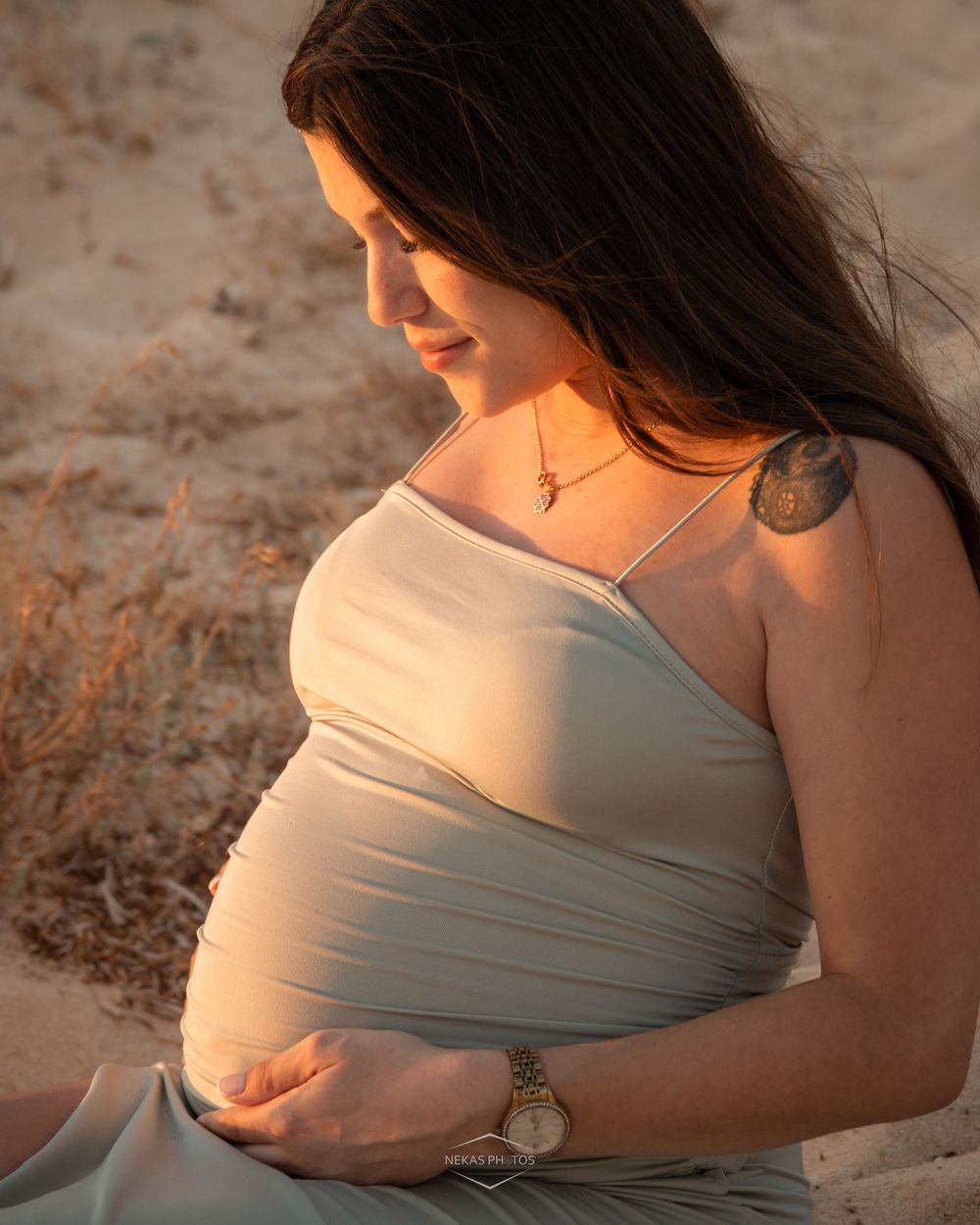 Pregnant woman in sage green dress cradling baby bump on sandy beach at golden hour sunset.