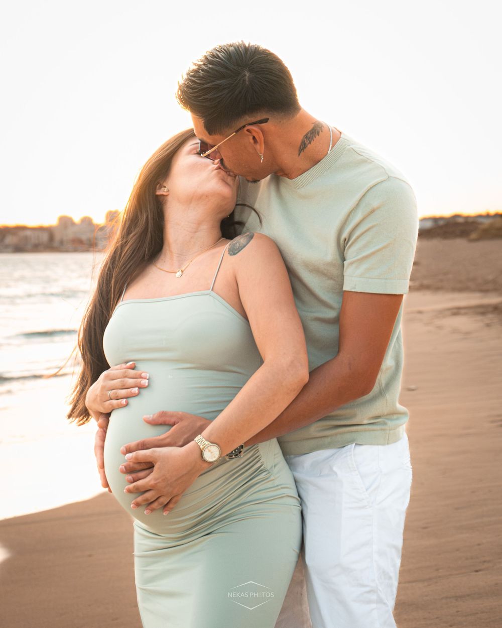 Couple sharing a kiss on the beach during golden hour, woman in mint dress cradling baby bump.