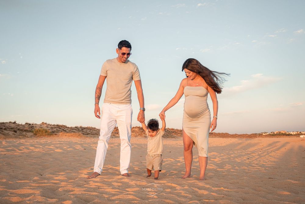 Pregnant family of three holding hands walking on sandy beach at golden hour sunset.