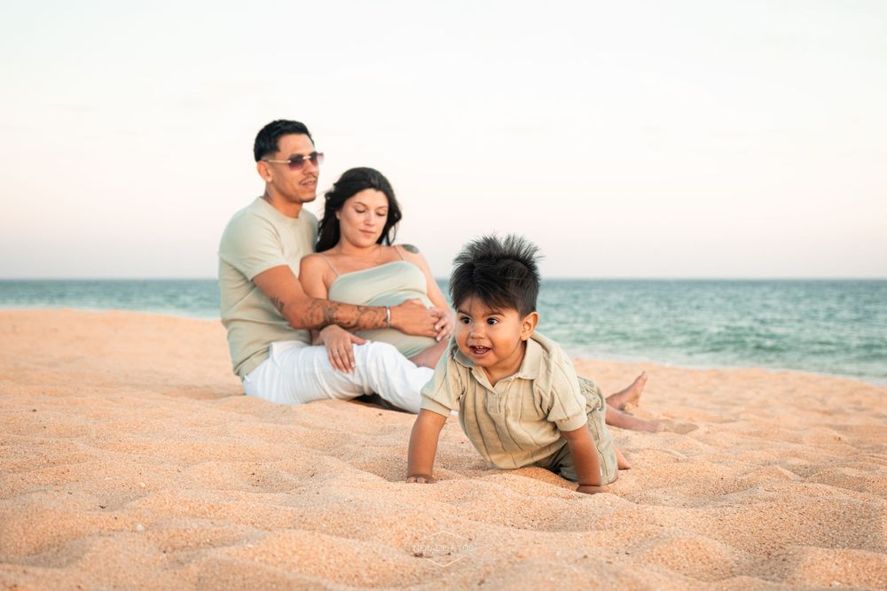 Family of three enjoying a sunny beach day, with parents relaxing on sand while toddler crawls in foreground.